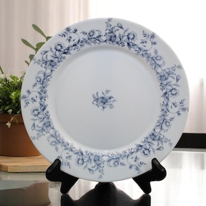 May include: White ceramic plate with a blue floral design around the rim and a small floral cluster in the center. The plate is displayed on a black stand, showcasing its decorative pattern. A classic piece for serving.