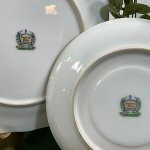 Op de afbeelding: Twee witte keramische borden met een gouden rand. De borden hebben een decoratief embleem in het midden met de tekst "Imperial China Made in Japan" en "KOMATSU" in groen. De borden zijn waarschijnlijk bedoeld om voedsel op te dienen.
