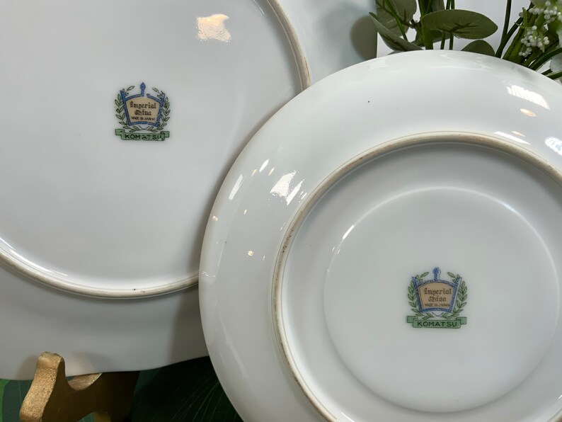 Op de afbeelding: Twee witte keramische borden met een gouden rand. De borden hebben een decoratief embleem in het midden met de tekst "Imperial China Made in Japan" en "KOMATSU" in groen. De borden zijn waarschijnlijk bedoeld om voedsel op te dienen.
