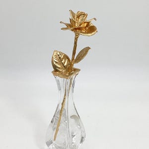 May include: A gold-colored rose sculpture with detailed petals and leaves, displayed in a clear, faceted glass vase. The rose stem extends into the vase, creating a decorative floral arrangement. The vase has a gold rim.