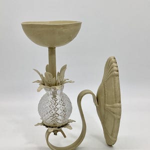 May include: A decorative wall sconce with a pineapple-shaped glass element and a beige finish. The sconce features a curved arm and a seashell-shaped backplate, adding a touch of elegance to any space. The sconce is designed to hold a candle or small light.