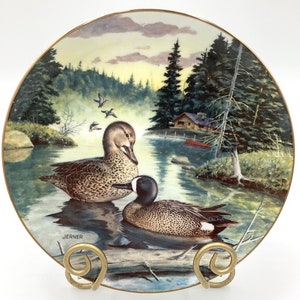 May include: A decorative ceramic plate featuring a painting of two blue-winged teal ducks in a lake setting. The plate has a gold rim and a signature "JERNER" in the bottom left corner.