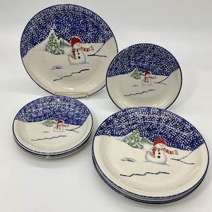 May include: A set of four ceramic plates with a winter theme. The plates feature a blue speckled border and a winter scene with a snowman wearing a red hat and scarf. The plates are white with a blue speckled border.