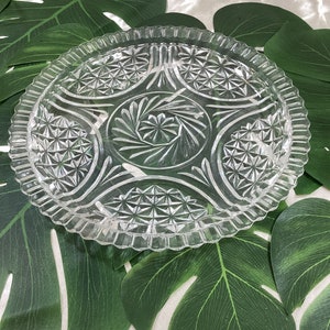 May include: A clear glass serving dish with a decorative, cut-glass design. The dish has a scalloped edge and a central, circular design.