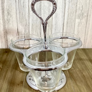 May include: A silver-toned serving piece with three clear glass cups. The cups are held in place by a decorative metal frame and a central handle. The base has a beaded edge.
