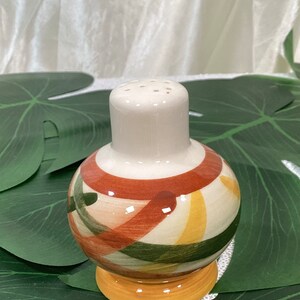 May include: A ceramic salt shaker with a cream-colored top and base. The spherical body features a pattern of orange, green, and yellow stripes. The shaker is designed with a classic, functional aesthetic.