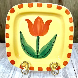 May include: A yellow ceramic square serving plate with an orange tulip design and a red and yellow border. The plate is on a gold metal stand.