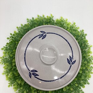 May include: A round, speckled gray ceramic lid with a dark blue leaf design. The lid has a small, round handle in the center and is surrounded by a green wreath. The leaf design is repeated in three places around the rim.