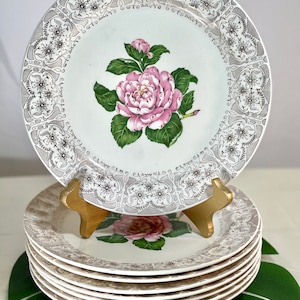May include: Set of eight white ceramic dinner plates with a delicate floral design. The plates feature a pink flower with green leaves and a silver-colored border with a repeating pattern.