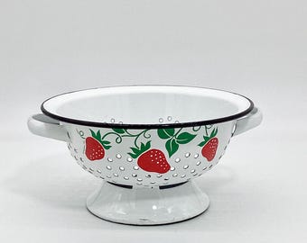 May include: White enamel colander with a black rim and a strawberry pattern. The colander has a pedestal base and two handles.