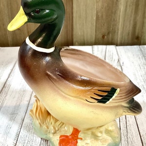 May include: A ceramic duck planter with a green head, yellow beak, and brown body. The duck is perched on a light green base with tan-colored grass details. The planter has a wide opening for plants or decorative items.