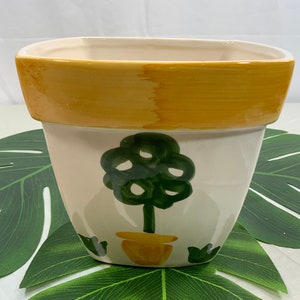 May include: A square ceramic planter with a cream-colored body and a yellow rim. A green tree design is painted on the front, with a yellow pot at the base. The planter is suitable for indoor or outdoor use.