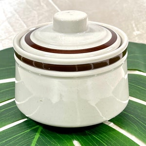 May include: A white ceramic sugar bowl with a lid. The bowl and lid are decorated with a brown band. The sugar bowl has a round shape and a small knob on top of the lid.