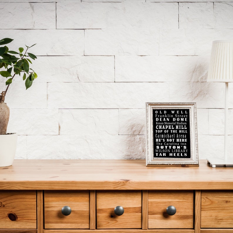 Op de afbeelding: Een ingelijst zwart-witdruk met de tekst "OLD WELL Franklin Street DEAN DOME Kenan Memorial Stadium CHAPEL HILL TOP OF THE HILL Carmichael Arena HE'S NOT HERE The Carolina Inn SUTTON'S WILSON LIBRARY TAR HEELS" op een houten plank met een plant in een pot en een lamp.