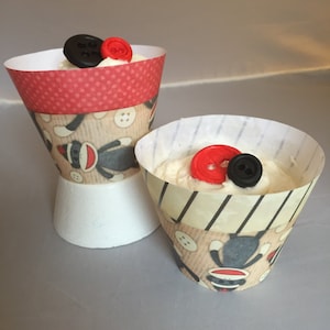 May include: Two cupcake wrappers with a sock monkey theme. One wrapper is red with white polka dots and the other is brown with white stripes. Both wrappers have red and black buttons on top.