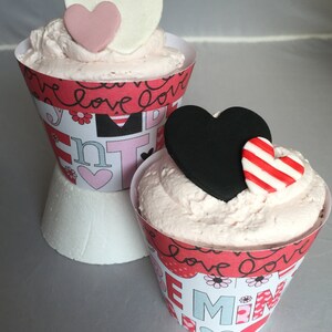 May include: Two cupcakes decorated with pink and black heart-shaped frosting and fondant. The cupcakes are in cone-shaped wrappers with a red, white, and black pattern and the words "love love love" and "remember me".