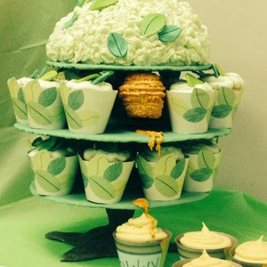 May include: A three-tiered cupcake stand with green and white cupcakes decorated with green leaves. The top tier has a large green and white cake with green leaves. The bottom tier has cupcakes with yellow frosting and the word "Sunny" written on one.