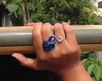Könnte beinhalten: Eine Hand trägt zwei Ringe. Der Ring am Zeigefinger ist ein silberner Knotenring. Der Ring am Mittelfinger ist ein blauer und violetter Drahtring mit einem Knotendesign.