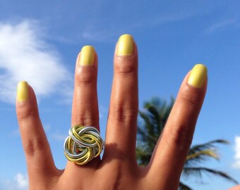 Könnte beinhalten: Eine Hand mit gelbem Nagellack trägt einen silbernen und goldenen Ring mit einem Knotendesign. Die Hand ist vor einem blauen Himmelhintergrund.
