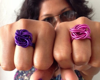 Könnte beinhalten: Zwei Nahaufnahmen von Händen, die Ringe tragen. Der Ring an der linken Hand ist lila und der Ring an der rechten Hand ist rosa. Beide Ringe sind aus gedrehtem Draht.