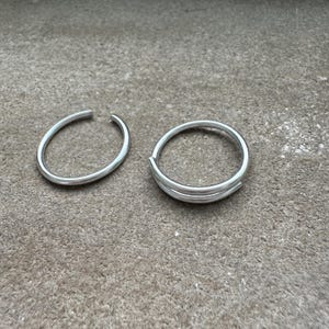 May include: Two silver rings are displayed on a gray surface. One ring is a simple open band. The other ring has a double-band design. The rings are made of a shiny, metallic material.
