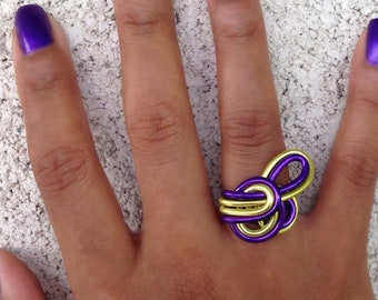 Könnte beinhalten: Ein goldener und violetter Drahtring mit einem einzigartigen Knotendesign. Der Ring wird am Mittelfinger einer Hand mit violetter Lackierung getragen.