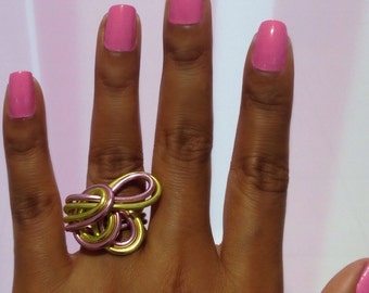 Könnte beinhalten: Eine Hand trägt einen Ring aus rosa und goldenem Draht. Der Ring besteht aus zwei ineinander verschlungenen Drähten, einem rosa und einem goldenen. Die Hand ist mit rosa Nagellack lackiert.