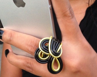 May include: A unique ring featuring a black and gold wire design. The ring is worn on a finger, with a silver smartphone in the background. The ring's design is abstract and eye-catching, with a mix of curved and looped wires.