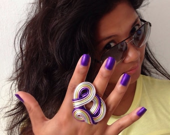 Könnte beinhalten: Nahaufnahme einer Frauenhand, die einen großen Ring aus silbernem und violettem Draht trägt. Der Ring besteht aus mehreren Drähten, die miteinander verdreht sind, um ein einzigartiges Design zu schaffen. Die Nägel der Frau sind in einem leuchtenden Violett lackiert.