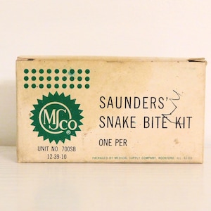 May include: A brown cardboard box with green and white text that reads "SAUNDERS' SNAKE BITE KIT" and "UNIT NO 700SB 12-39-10". The box also has a green logo with the letters "MJCo" inside a circle.