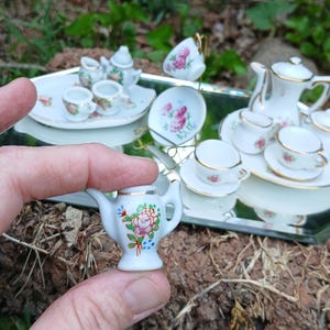 May include: A miniature white porcelain teapot featuring a hand-painted floral design with a pink rose, green leaves, and blue accents, finished with a gold rim. The teapot has a curved handle and spout, and is held between two fingers.