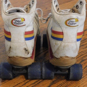 May include: A pair of white roller skates with blue, red, and yellow stripes. The skates have a Free Former logo on the back of each boot. The wheels are black with blue hubs.
