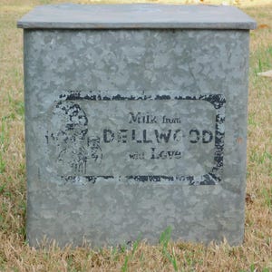 May include: A galvanized metal container with a lid, likely for storage. The container has a rectangular shape and a faded label that reads "Milk from DELLWOOD with Love."