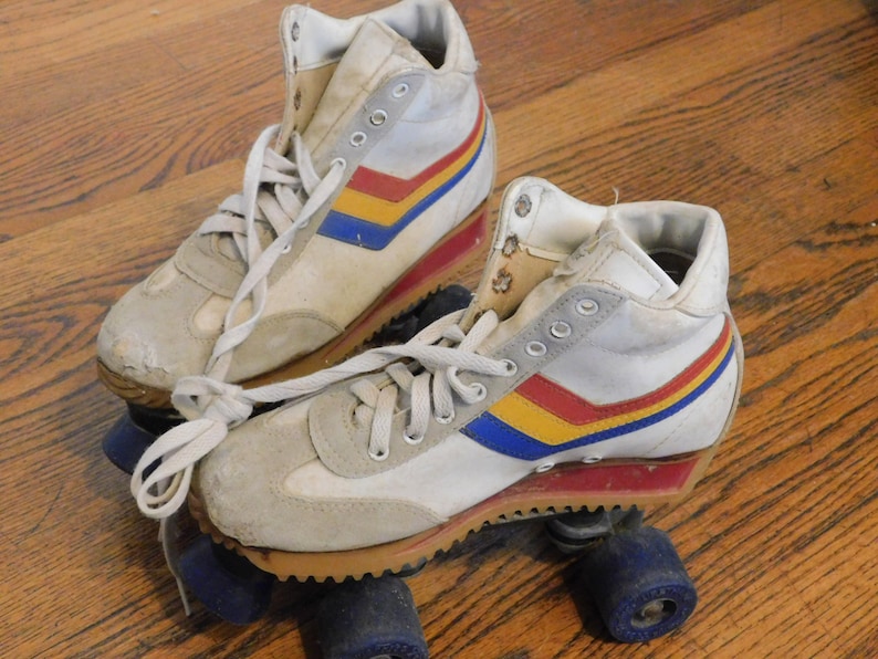 May include: A pair of white roller skates with blue, yellow, and red stripes. The skates have white laces and blue wheels.