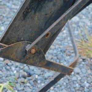K&ouml;nnte beinhalten: Nahaufnahme eines rostigen Metallgelenks mit einem Bolzen und einer Mutter. Das Gelenk ist an einem Metallrahmen befestigt. Der Hintergrund ist eine Schotterfl&auml;che.