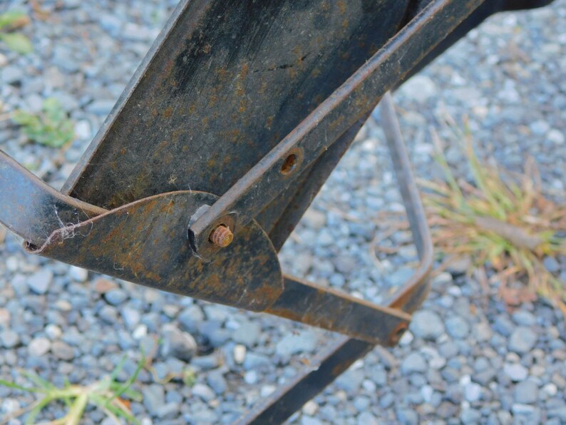 K&ouml;nnte beinhalten: Nahaufnahme eines rostigen Metallgelenks mit einem Bolzen und einer Mutter. Das Gelenk ist an einem Metallrahmen befestigt. Der Hintergrund ist eine Schotterfl&auml;che.