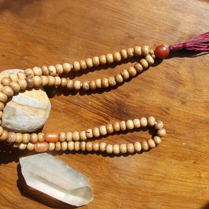 May include: A wooden mala bead necklace with a red tassel and a clear quartz crystal. The mala beads are brown and have a natural wood finish. The tassel is made of a soft, red fabric. The quartz crystal is clear and has a smooth, polished surface.