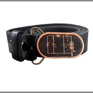 Puede incluir: Un cinturón de cuero negro con una hebilla de cobre que presenta un diagrama de circuito. La hebilla tiene un fondo negro con un diagrama de circuito de color cobre.