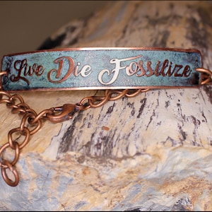 May include: A copper-colored bracelet with the words "Live Die Fossilize" in a cursive font. The rectangular centerpiece has a turquoise patina. The bracelet is connected by a chain with a lobster clasp.