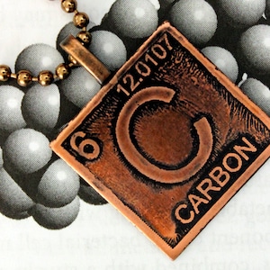 Może przedstawiać: Naszyjnik z miedzianym wisiorem w kształcie kwadratu przedstawiającym pierwiastek węgiel. Wisiorek ma liczbę atomową 6, masę atomową 12,0107 i literę C. Słowo "WĘGIEL" jest napisane na wisiorku.