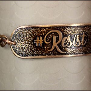 May include: A copper-colored bracelet with the word "Resist" in elegant script. The bracelet features a textured, rectangular plate with the word "Resist" and a hashtag symbol. The bracelet is attached to a chain.