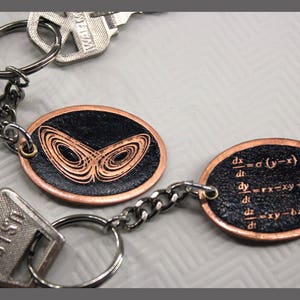 May include: A copper keychain with two black leather oval charms. One charm features a copper-colored butterfly design and the other charm has a black background with white mathematical equations. The keychain is attached to a silver key ring.