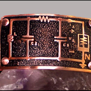 May include: A red leather bracelet with a copper plate featuring a circuit diagram. The diagram includes components like resistors, capacitors, and a transistor. The plate has a textured surface and is secured to the leather band with two rivets.