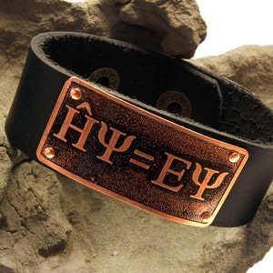 Könnte beinhalten: Ein schwarzes Lederarmband mit einer kupfernen Platte, die mit den griechischen Buchstaben "ΗΨ=ΕΨ" graviert ist.