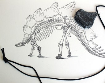 May include: A black and white illustration of a Stegosaurus skeleton with a black stone pendant on a black cord. The pendant is dark gray and has a textured surface. The dinosaur drawing is detailed.