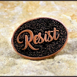 May include: An oval-shaped pin with the word "Resist" in copper-colored script. The background is black with a textured appearance, and the pin has a copper-colored border. The pin is set against a light-colored, textured surface.