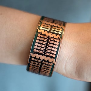 Puede incluir: Brazalete de cobre con diseño geométrico. El brazalete presenta una serie de formas y líneas conectadas en un patrón repetitivo. El cobre tiene un tono marrón rojizo cálido, con acentos verde oscuro. El brazalete mide aproximadamente 3,8 cm de ancho.