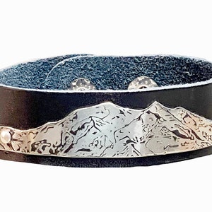 May include: Black leather cuff bracelet with a silver mountain range design. The bracelet has a snap closure and is approximately 1.5 inches wide. The mountain range is etched into the silver.