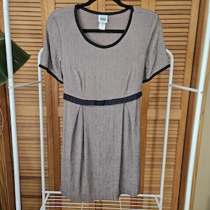 May include: A short-sleeved dress featuring a brown and black micro-check pattern, a round neckline with black trim, and a black horizontal band at the waist. The dress has a label at the neck that reads "KAREN KANE" and a straight skirt silhouette.