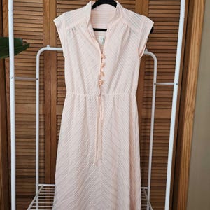 May include: A pale pink knee-length dress featuring a mandarin collar, cap sleeves, and a vertical row of small fabric-covered buttons down the center. The fabric has a subtle diagonal striped texture and an elasticized waist with a drawstring.